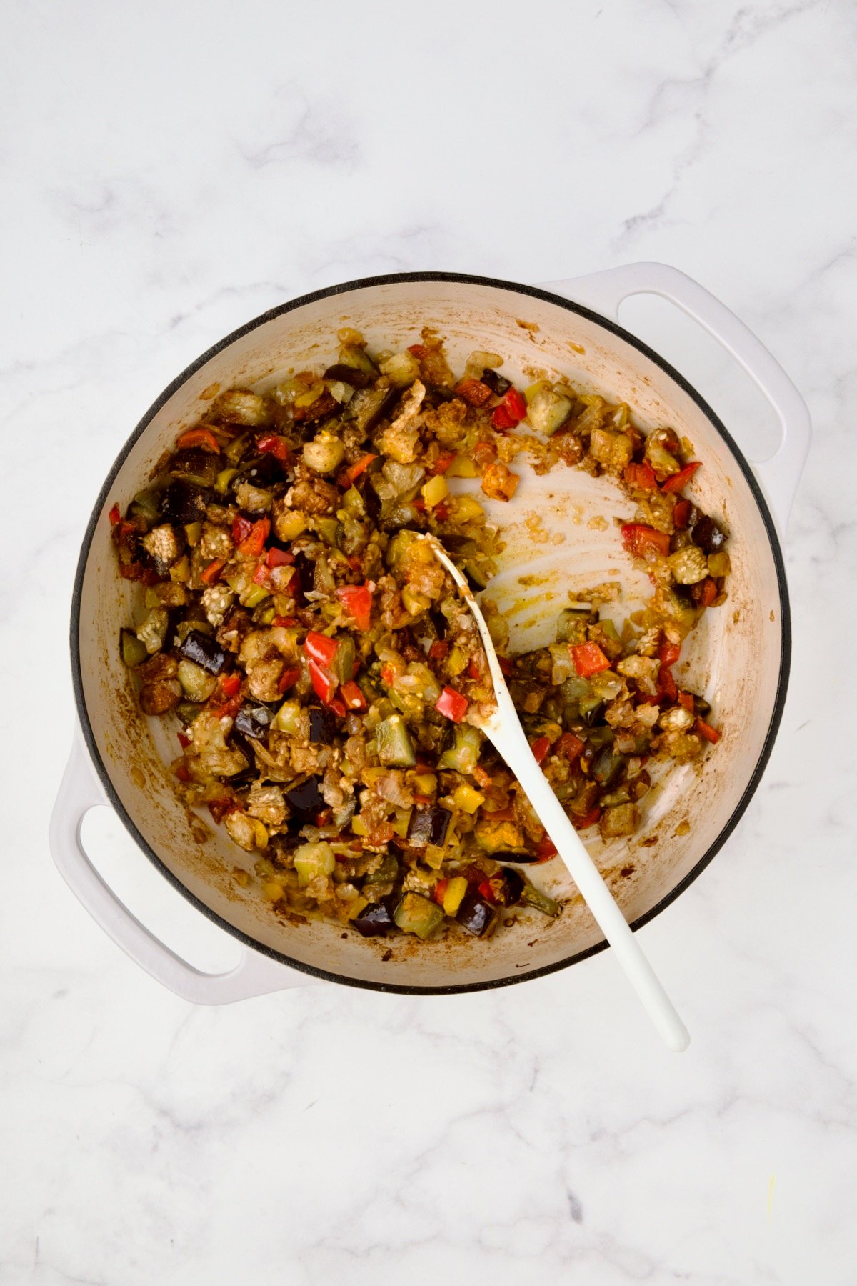 Cooked yellow bell peppers, red bell peppers, eggplant, and onions in white enamel cast iron skillet with garlic and spices added and stirred into vegetables.