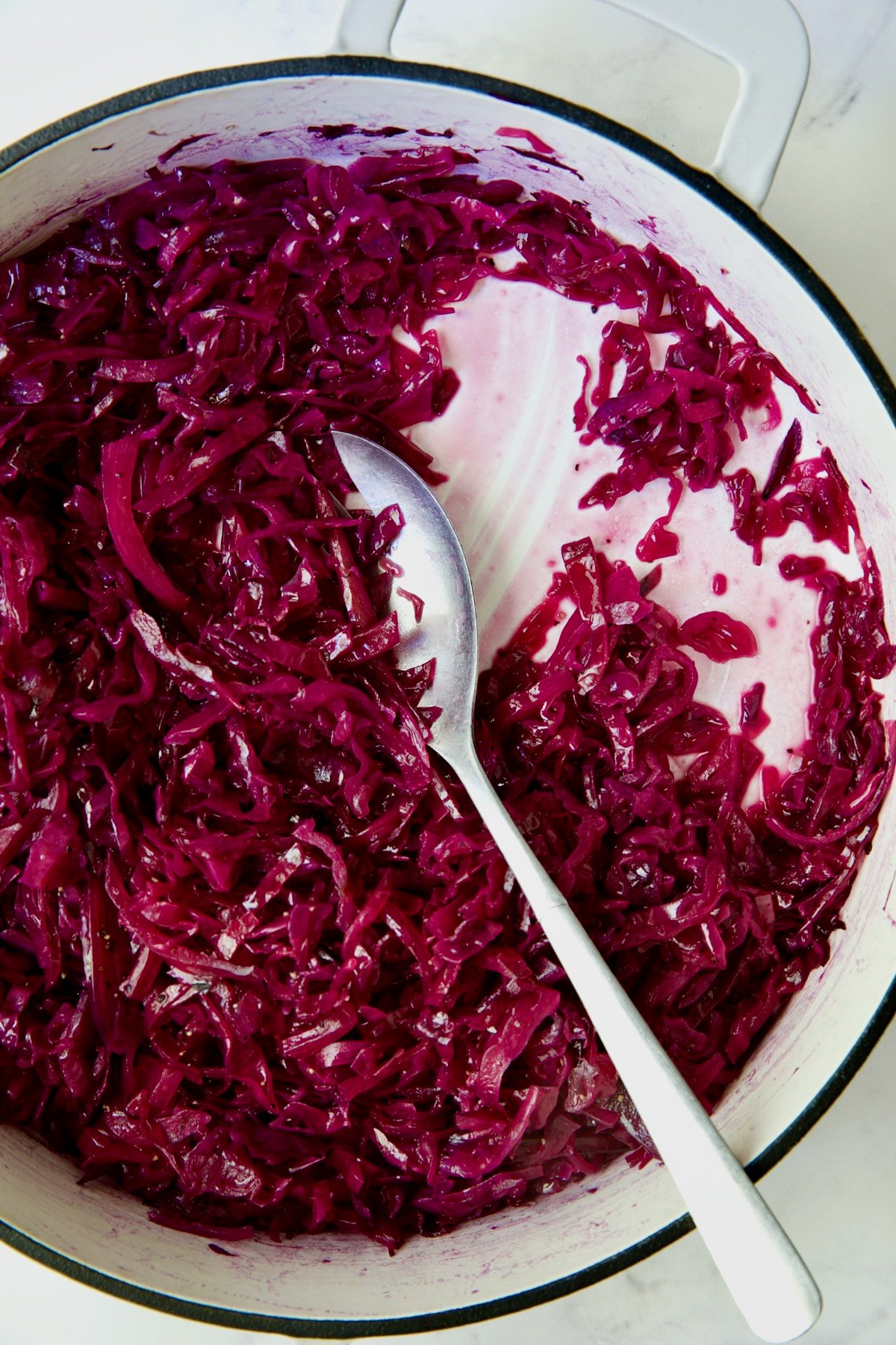 Fully cooked red cabbage in white enamel cast iron skillet with serving spoon.
