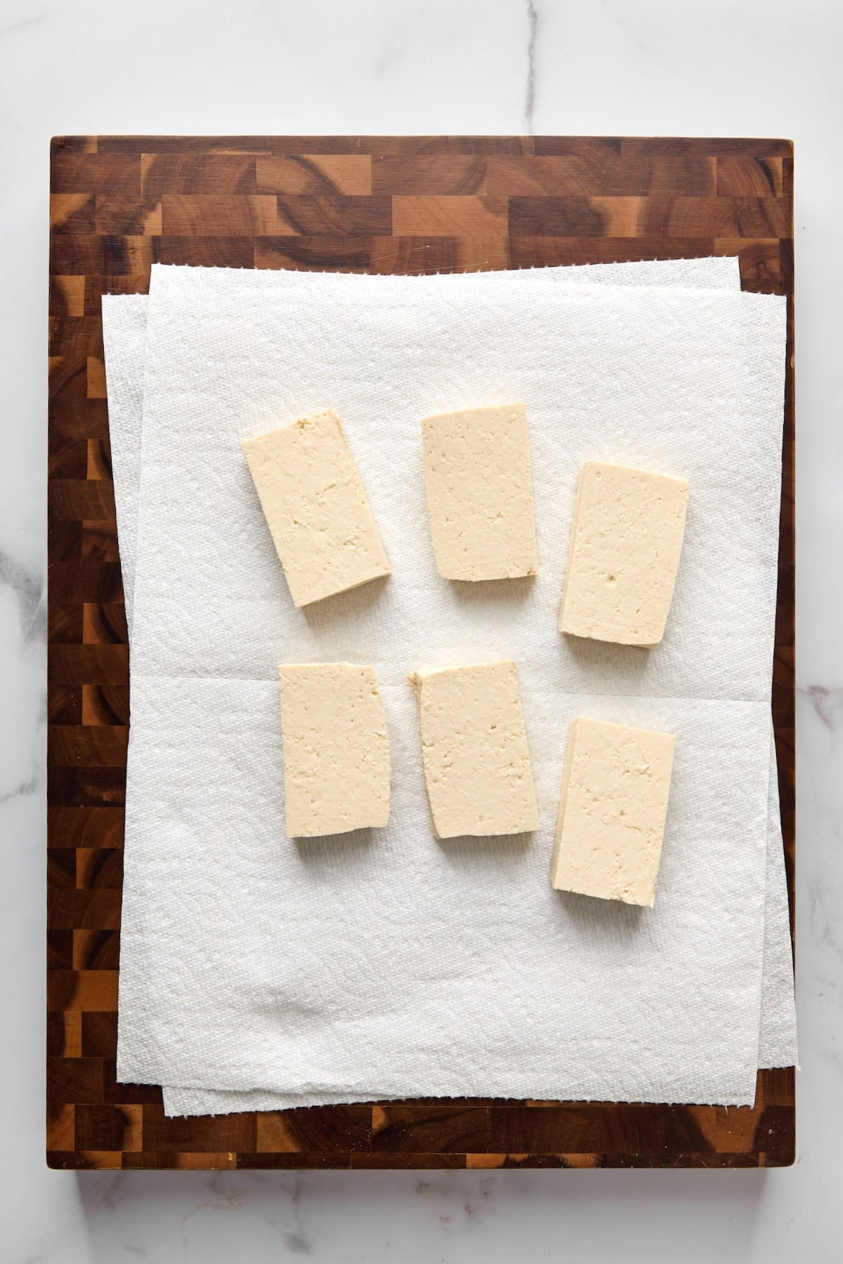 Six pieces of tofu on paper towels on wood cutting board.