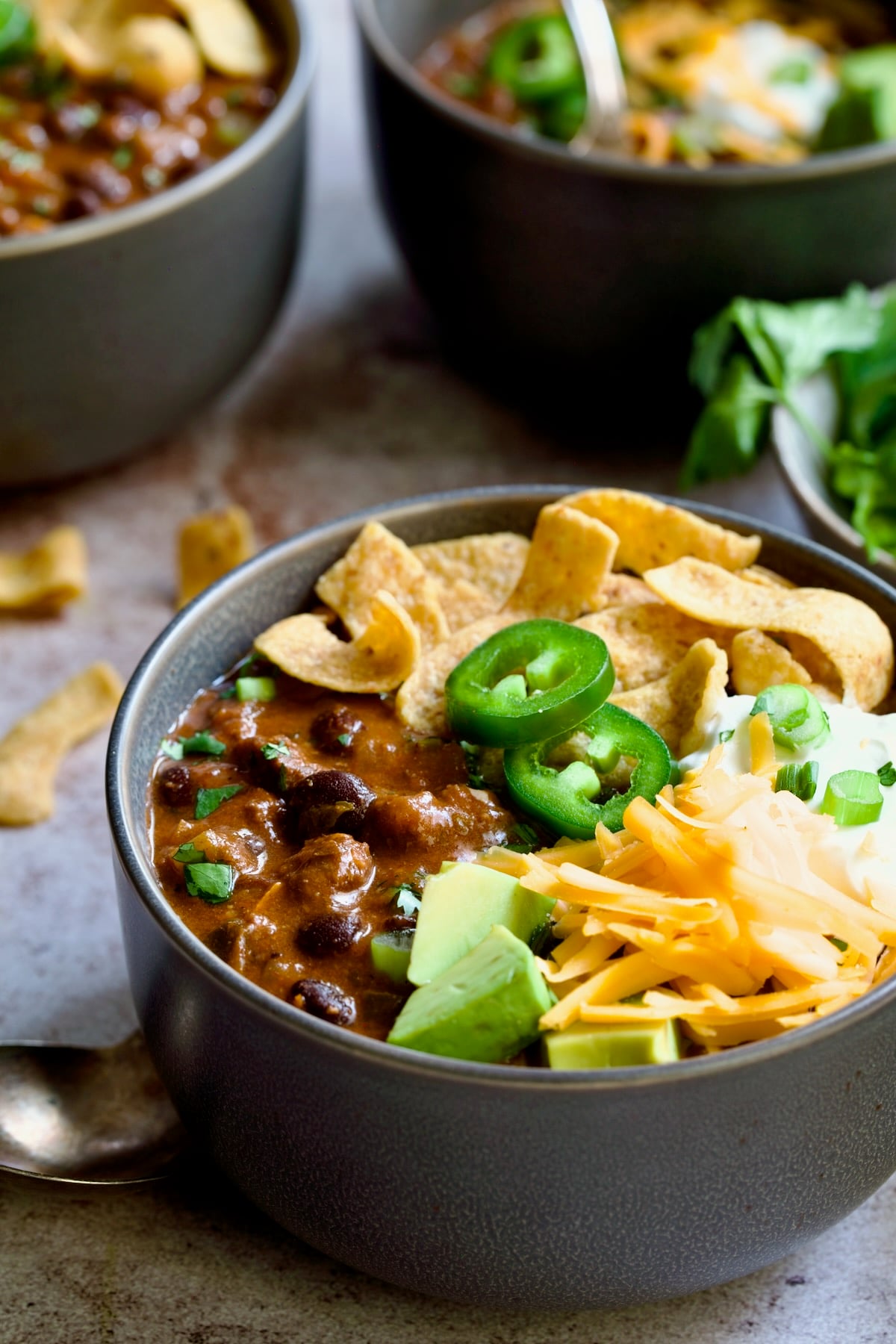 One serving of Beef and Chorizo Chili in gray bowl garnished with avocado, cheddar cheese, sour cream, jalapeno slices and corn chips.