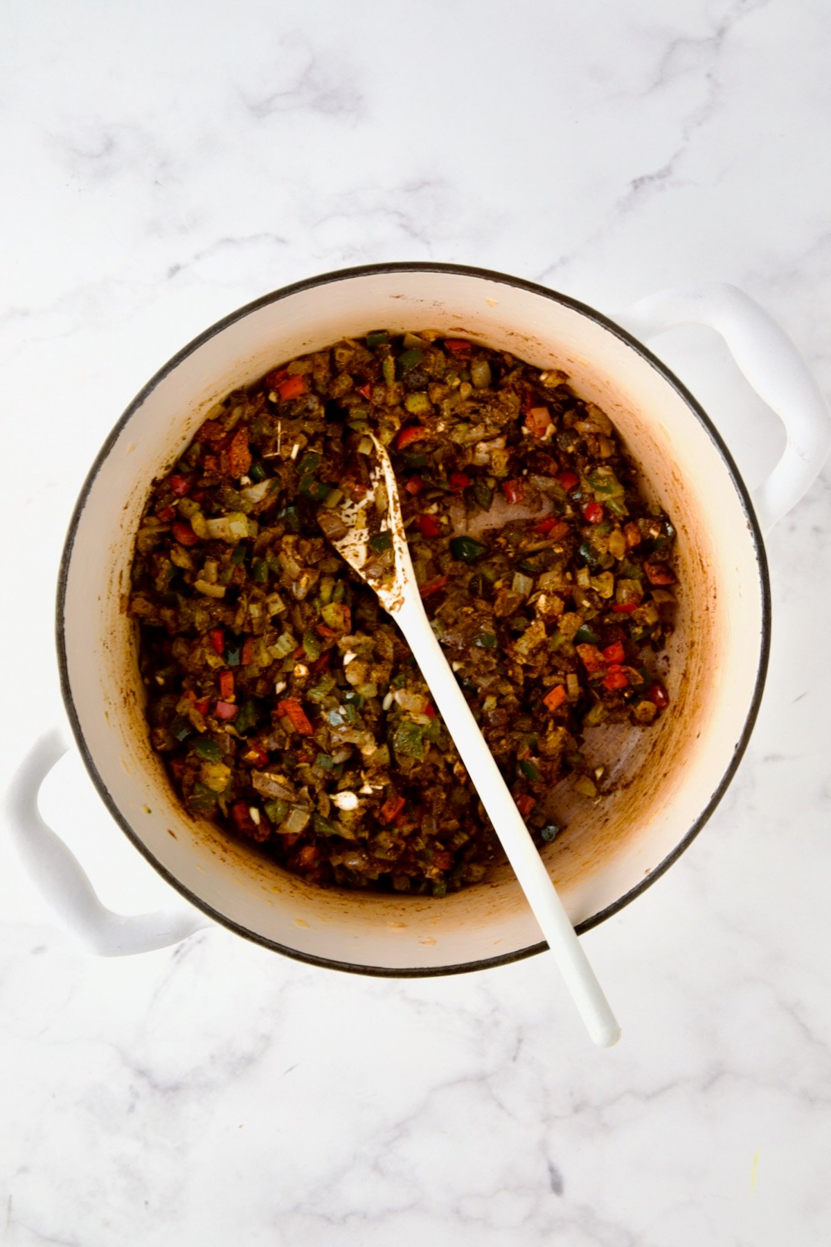 Spices stirred into cooked onions, celery, and peppers in white enamel Dutch oven with white mixing spoon.
