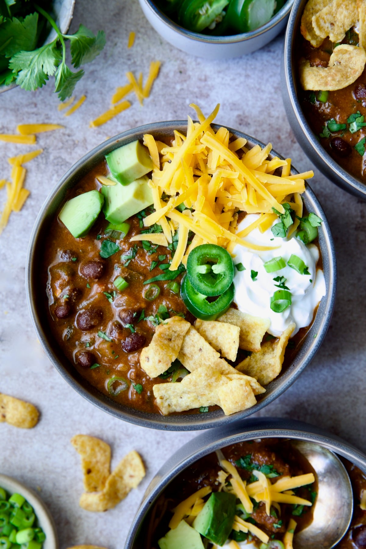 One serving of Beef and Chorizo Chili in gray bowl garnished with avocado, cheddar cheese, sour cream, jalapeno slices and corn chips.