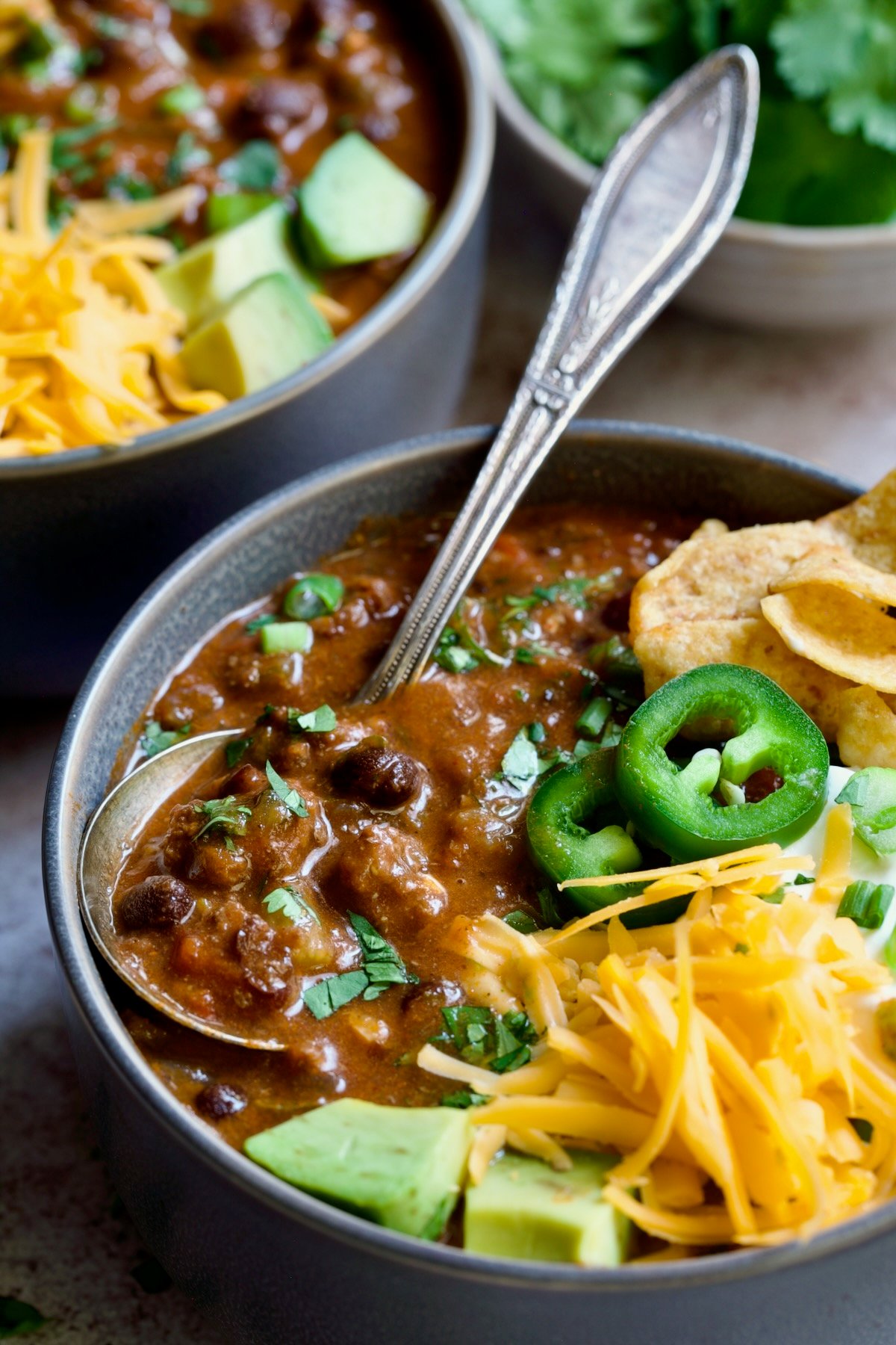 One serving of Beef and Chorizo Chili in gray bowl garnished with avocado, cheddar cheese, sour cream, jalapeno slices and corn chips.