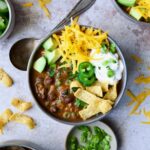 One serving of Beef and Chorizo Chili in gray bowl garnished with avocado, cheddar cheese, sour cream, jalapeno slices and corn chips.