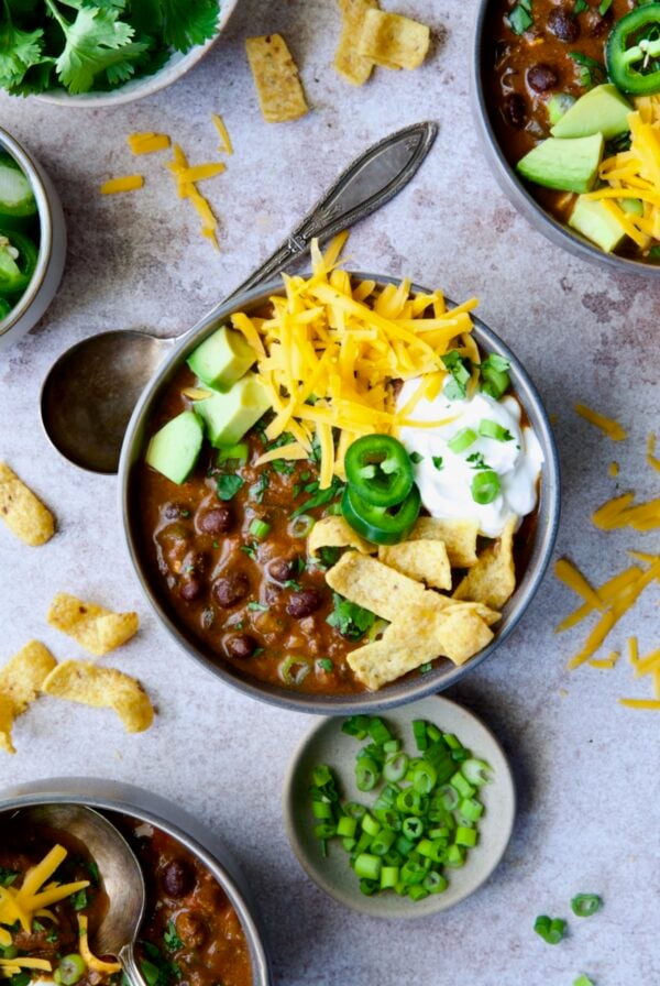 One serving of Beef and Chorizo Chili in gray bowl garnished with avocado, cheddar cheese, sour cream, jalapeno slices and corn chips.