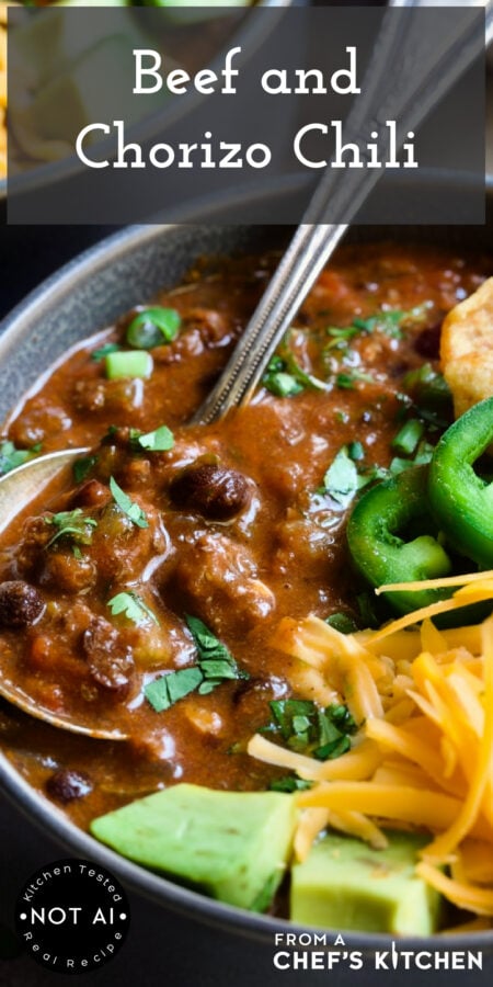 Pinterest graphic for Beef and Chorizo Chili showing one serving in a gray bowl garnished with avocado, cheddar, jalapeno, corn chips and sour cream.