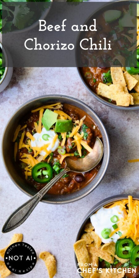 Pinterest graphic for Beef and Chorizo Chili showing three servings in a gray bowl garnished with avocado, cheddar, jalapeno, corn chips and sour cream.