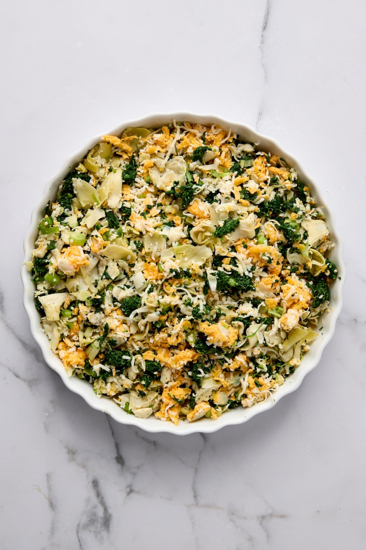 Ingredients for quiche in white quiche pan after more cheese is added and befpre custard are added.