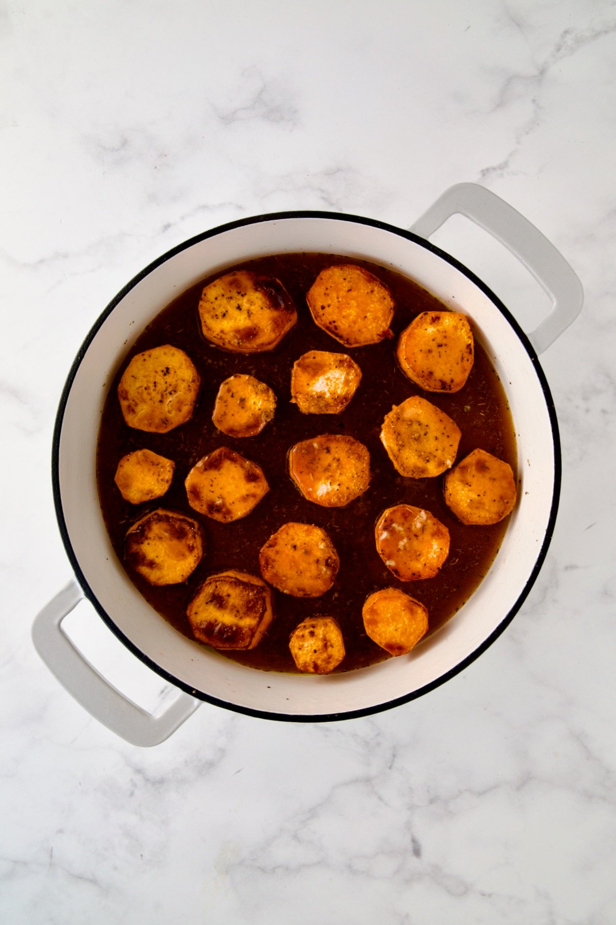Rounds of browned sweet potatoes in white enamel cast iron skillet in herb and broth blend.