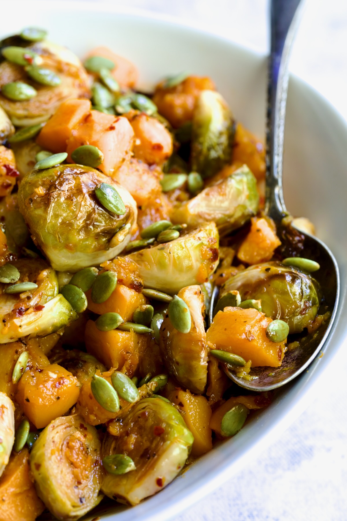 Maple Chili Crisp Brussels Sprouts and Butternut Squash garnished with pepitas in white bowl with serving spoon.