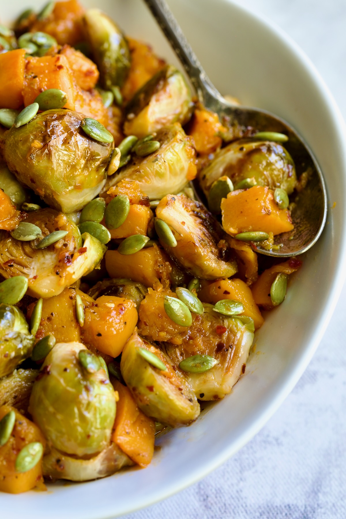 Maple Chili Crisp Brussels Sprouts and Butternut Squash garnished with pepitas in white bowl with serving spoon.