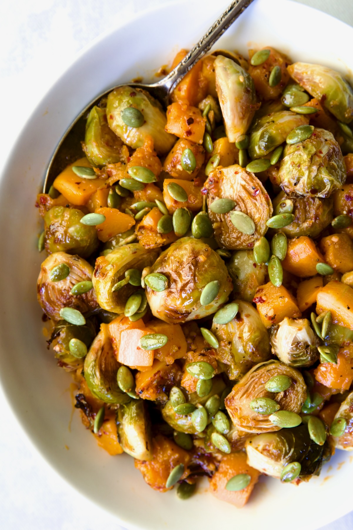Maple Chili Crisp Brussels Sprouts and Butternut Squash garnished with pepitas in white bowl with serving spoon.
