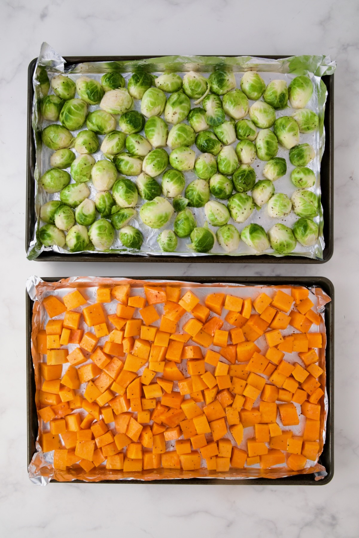 Halved Brussels sprouts and cubed butternut squash on nonstick aluminum foil lined rimmed baking sheets before being roasted.