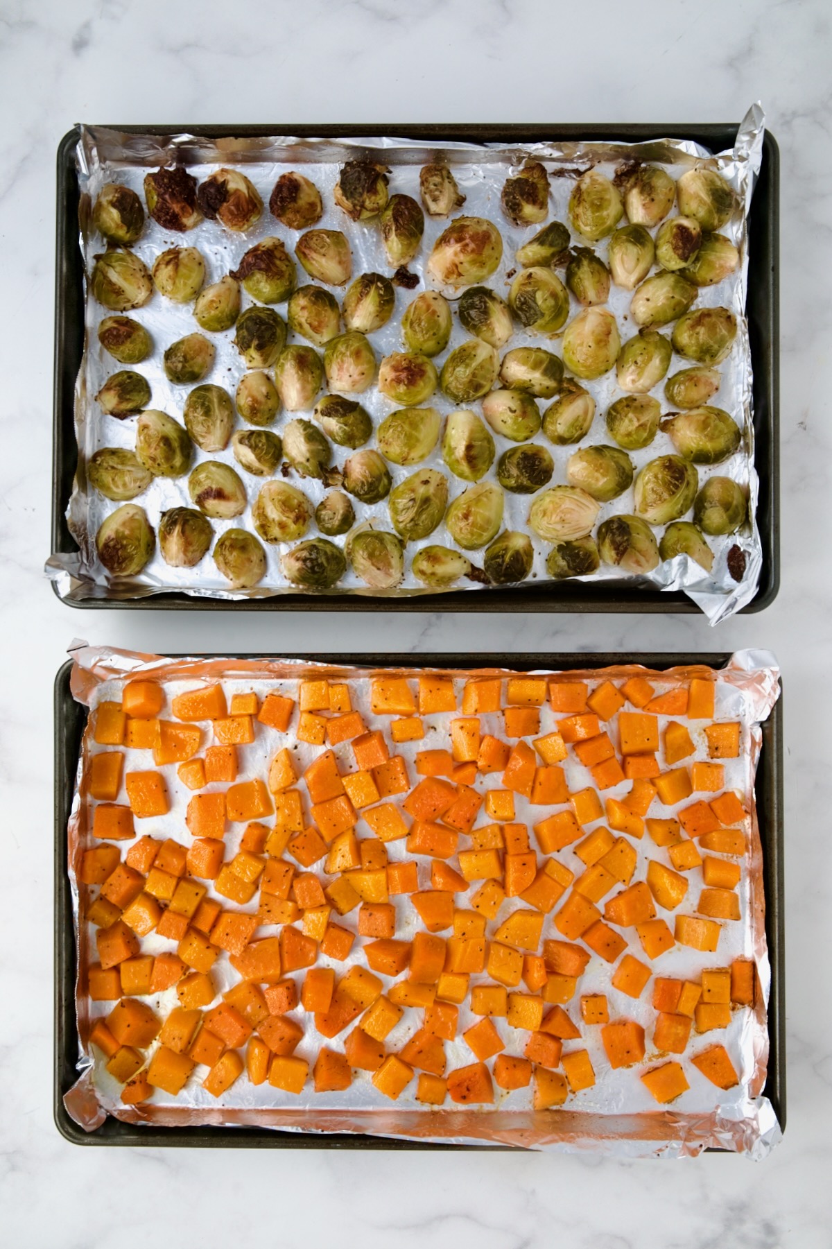 Halved Brussels sprouts and cubed butternut squash on nonstick aluminum foil lined rimmed baking sheets after being roasted.