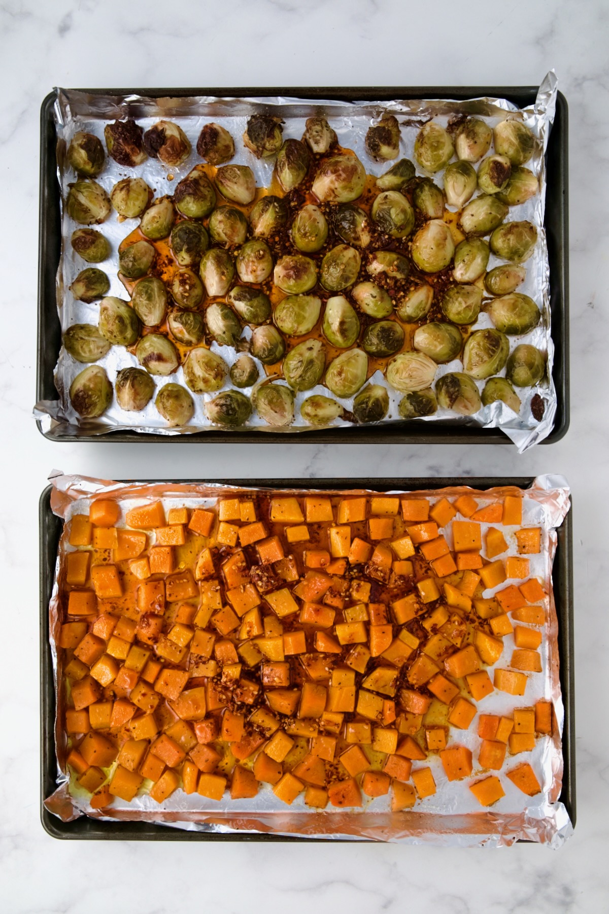 Halved Brussels sprouts and cubed butternut squash on nonstick aluminum foil lined rimmed baking sheets after being roasted with the sauce poured over.