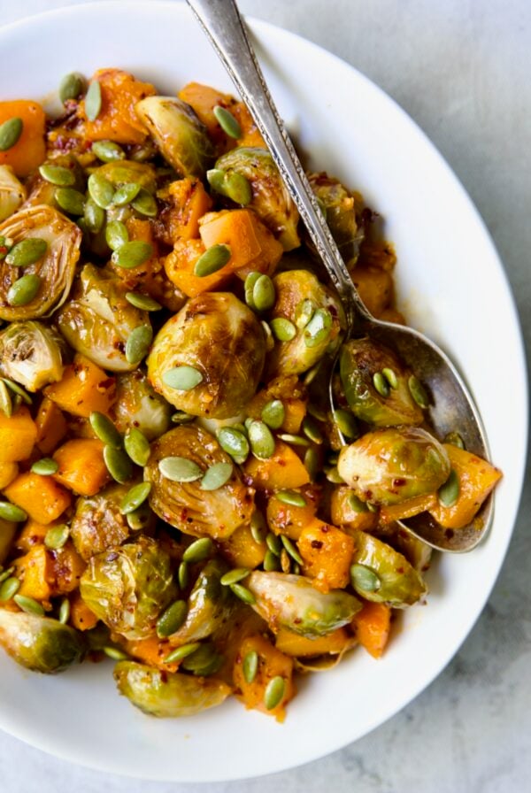 Maple Chili Crisp Brussels Sprouts and Butternut Squash garnished with pepitas in white bowl with serving spoon.