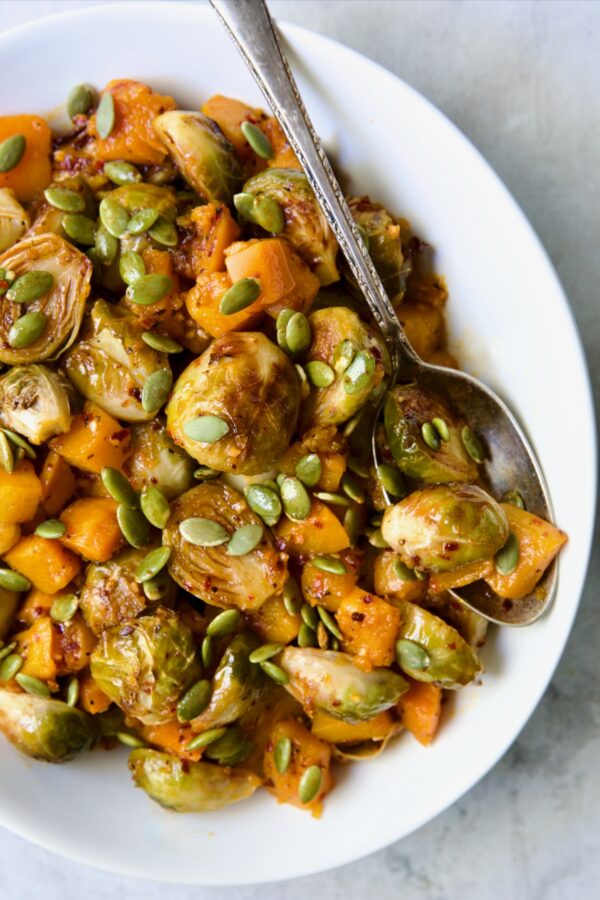 Maple Chili Crisp Brussels Sprouts and Butternut Squash garnished with pepitas in white bowl with serving spoon.