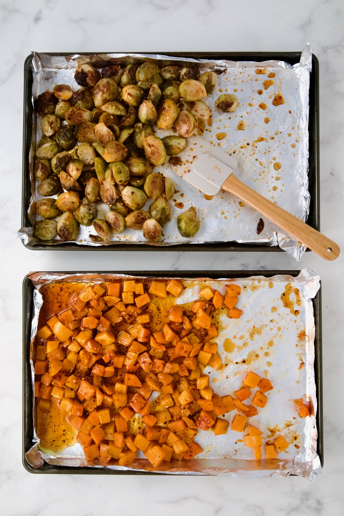 Halved Brussels sprouts and cubed butternut squash on nonstick aluminum foil lined rimmed baking sheets after being roasted with a wooden spatula stirring them.