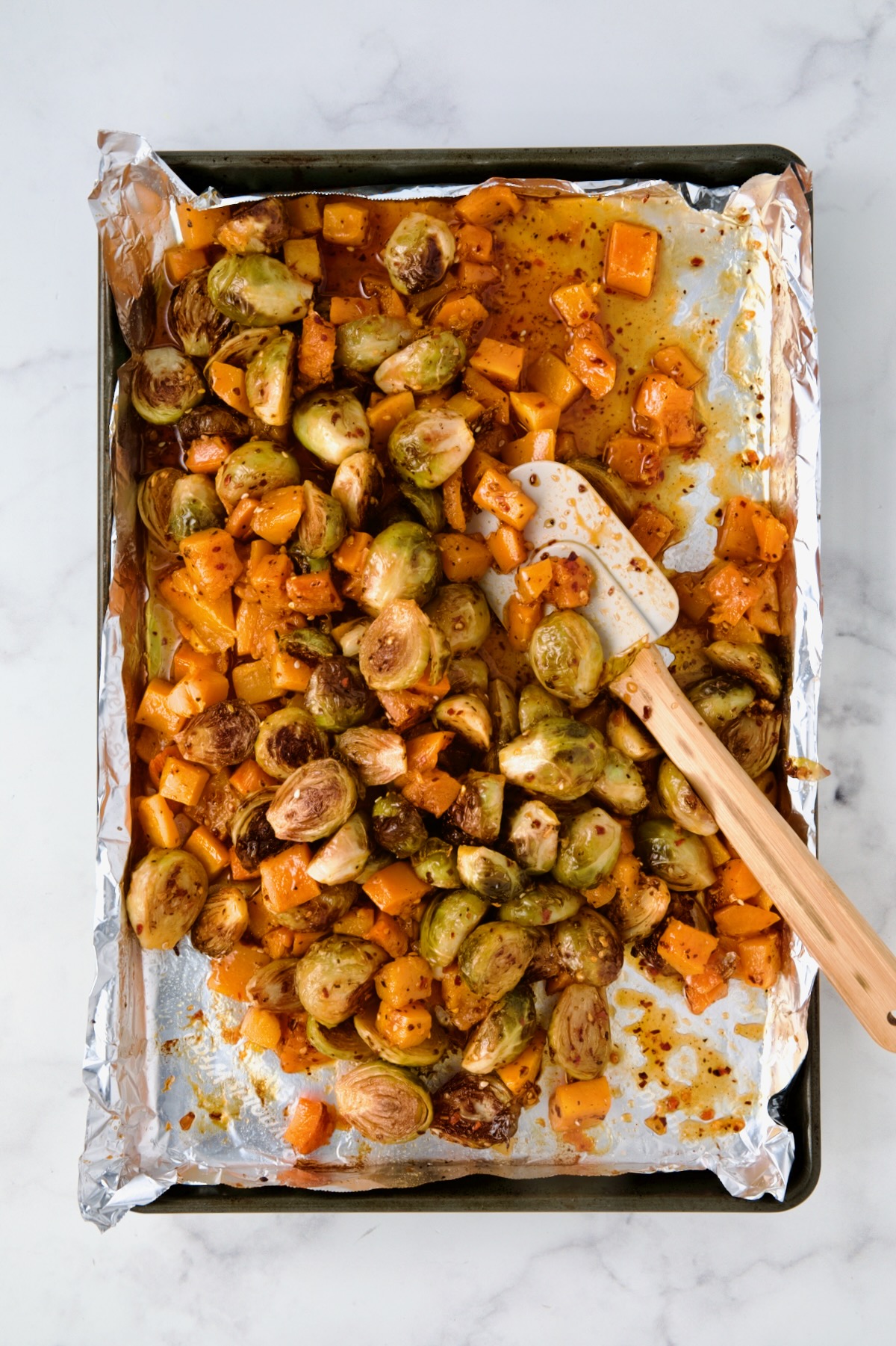 Roasted Brussels Sprouts and butternut squash being stirred with wooden spatula on foil-lined rimmed baking sheet.