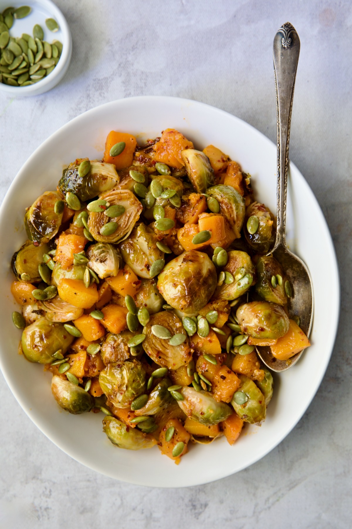 Maple Chili Crisp Brussels Sprouts and Butternut Squash garnished with pepitas in white bowl with serving spoon.