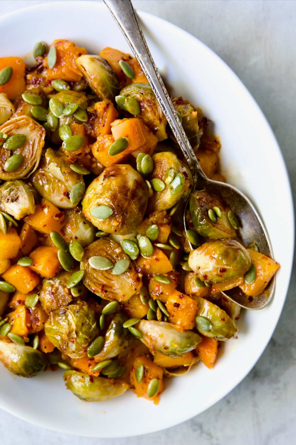 Maple Chili Crisp Brussels Sprouts and Butternut Squash garnished with pepitas in white bowl with serving spoon.