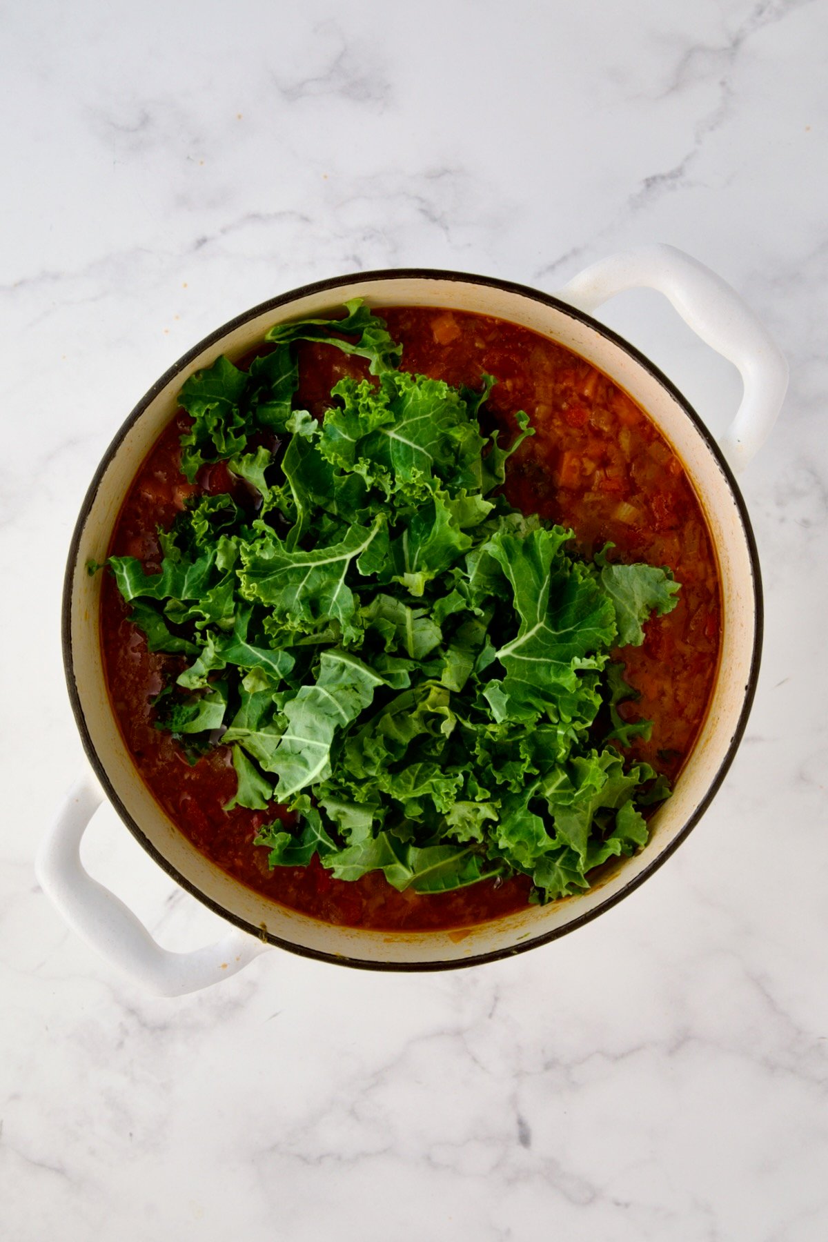 Kale added to soup in white enamel Dutch oven.