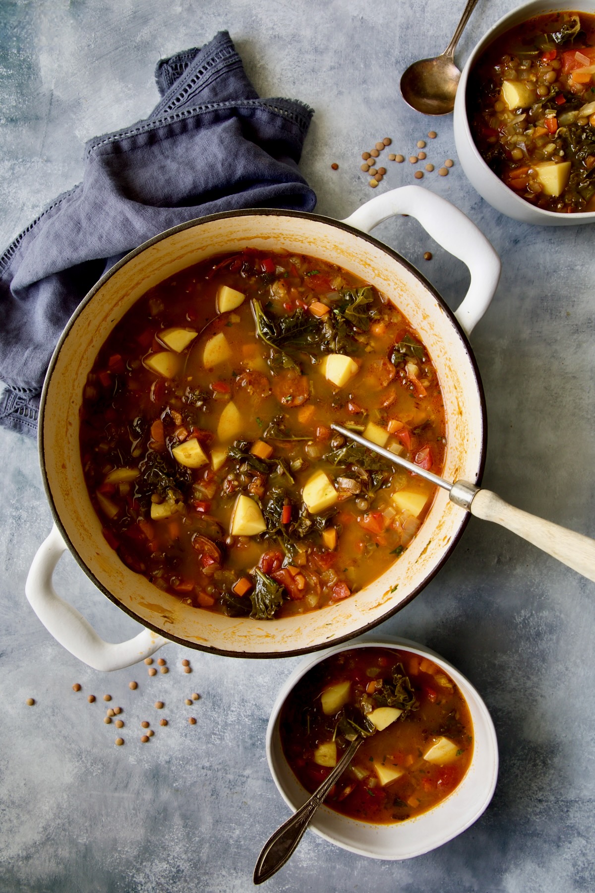 Finished Spanish Lentil Potato Soup in white enamel Dutch oven with soup ladle with some served up in white soup bowls.