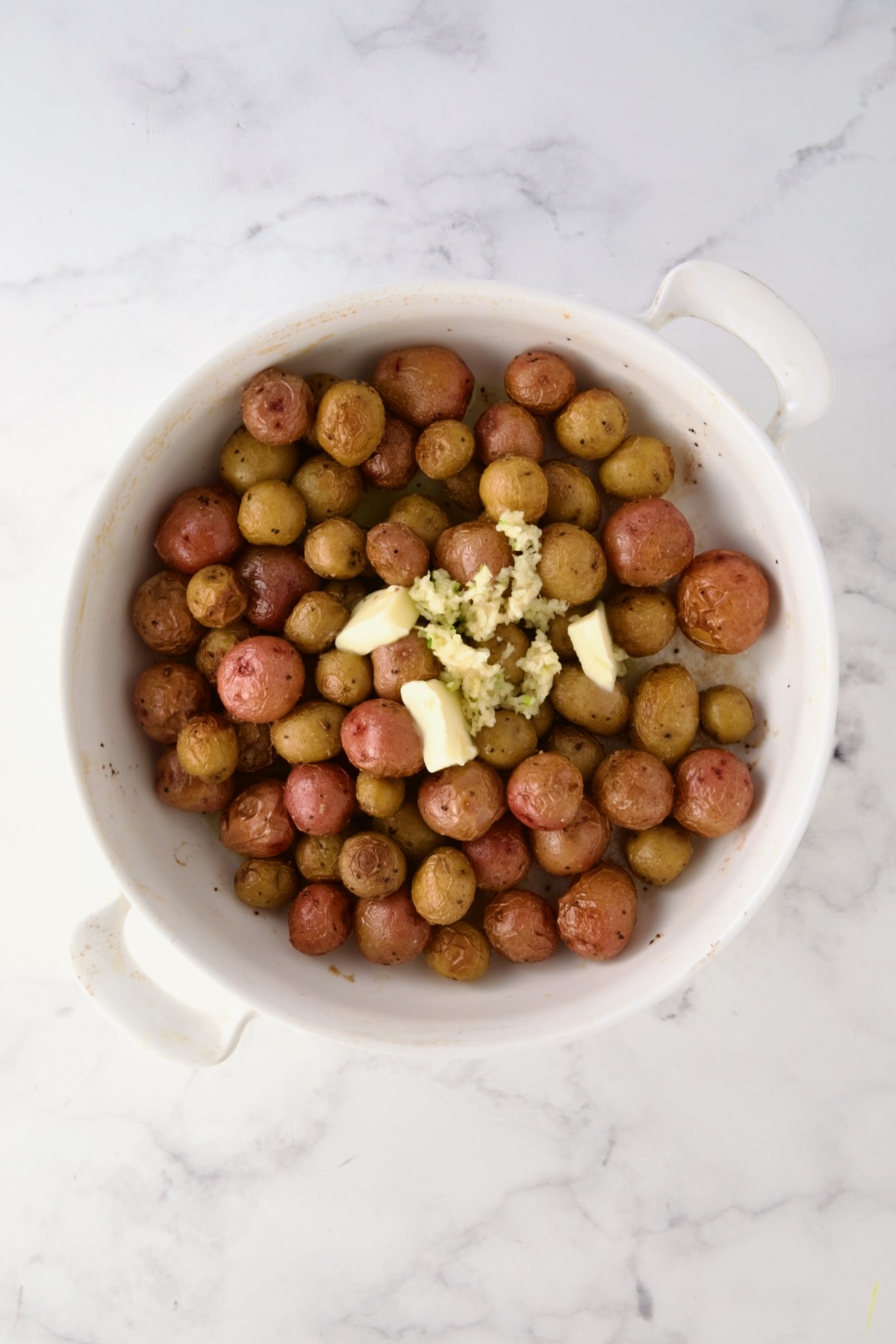 Butter and garlic added to roasted mini potatoes in round white baking dish.