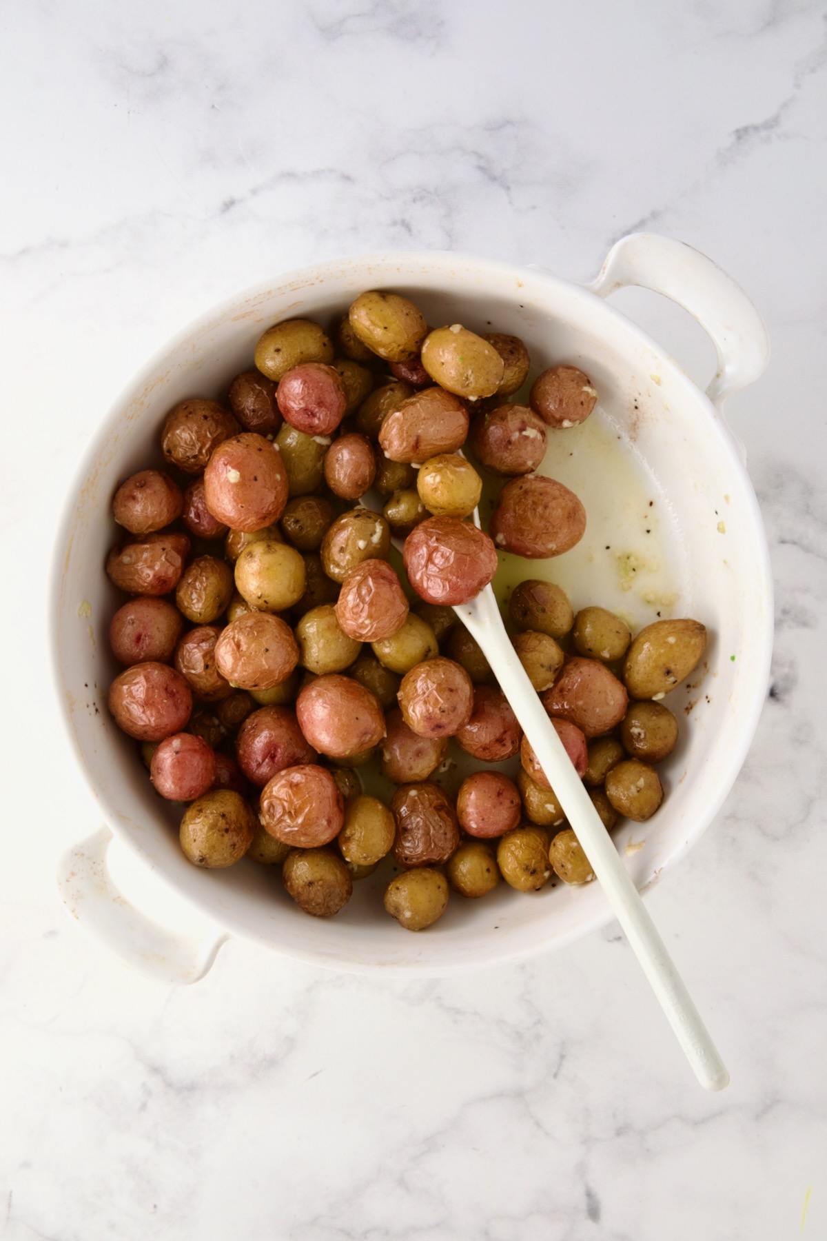 White mixing spoon stirring butter and garlic into mini potatoes in round white baking dish.