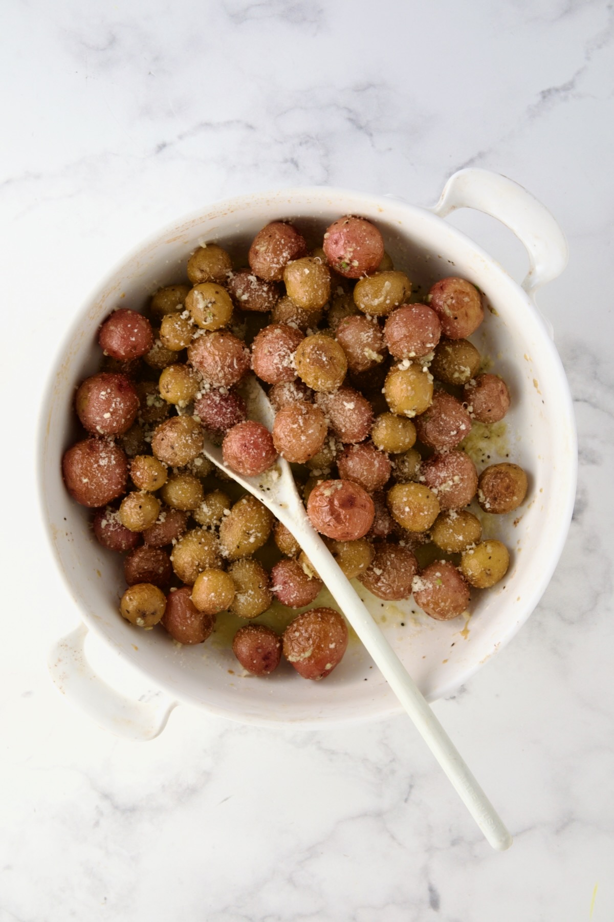 Parmesan cheese added and being stirred into to mini potatoes in round white baking dish with white mixing spoon.