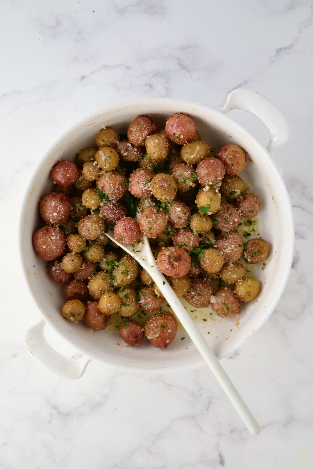 Parsley added to roasted mini potatoes and being stirred with white mixing spoon in round white baking dish.