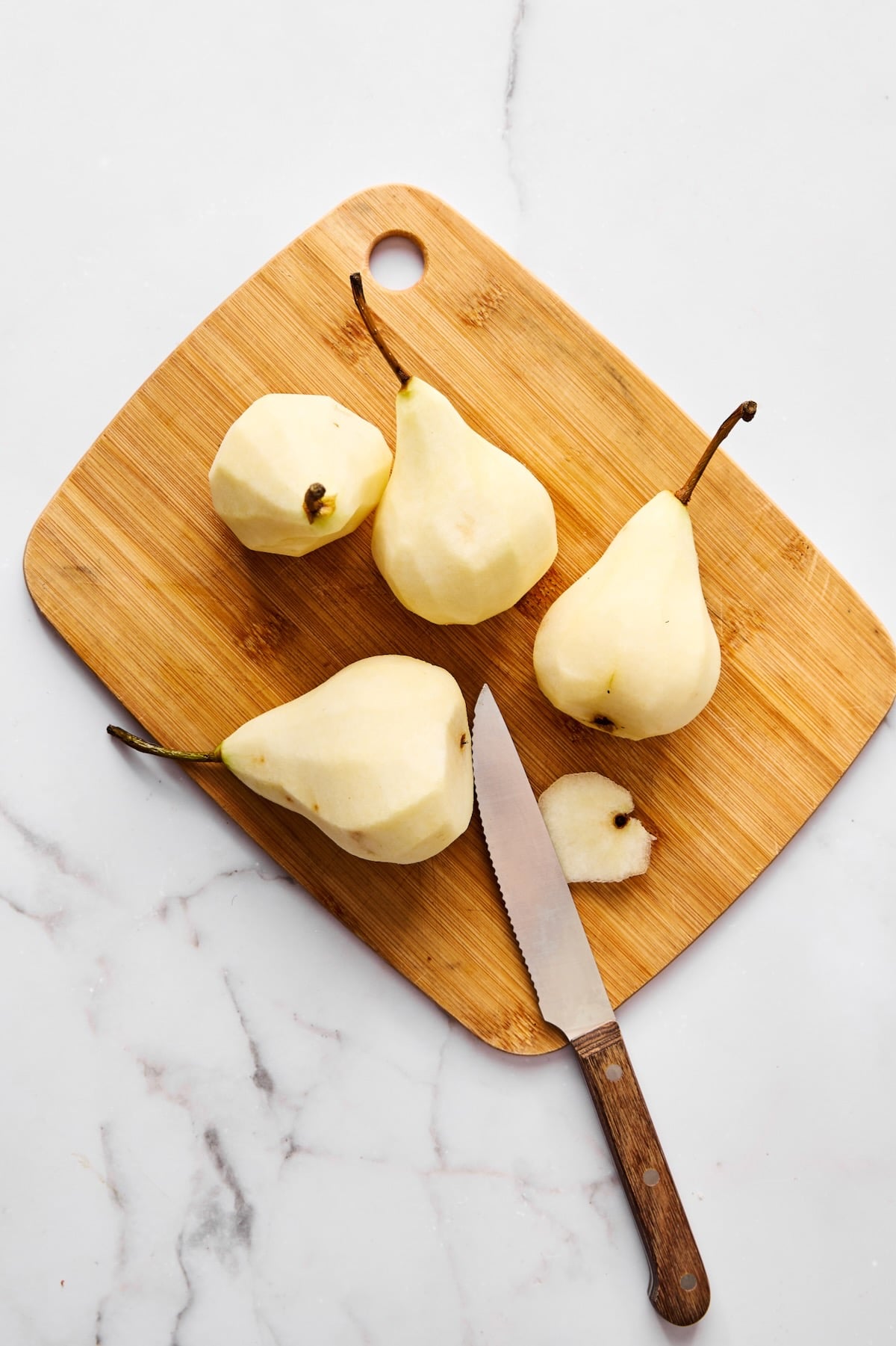 Four peeled pears with knife on wood cutting board.