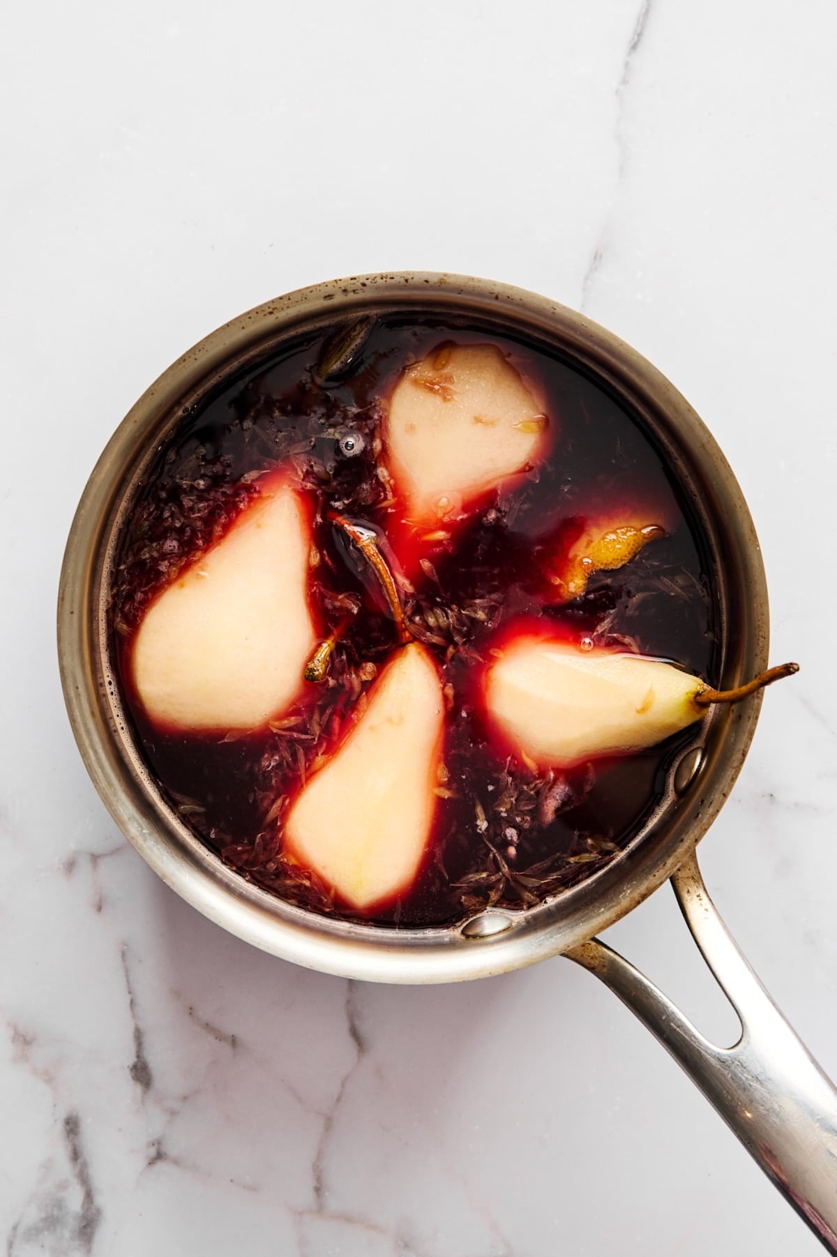 Four pears in stainless steel saucepan being simmered in red wine sauce.