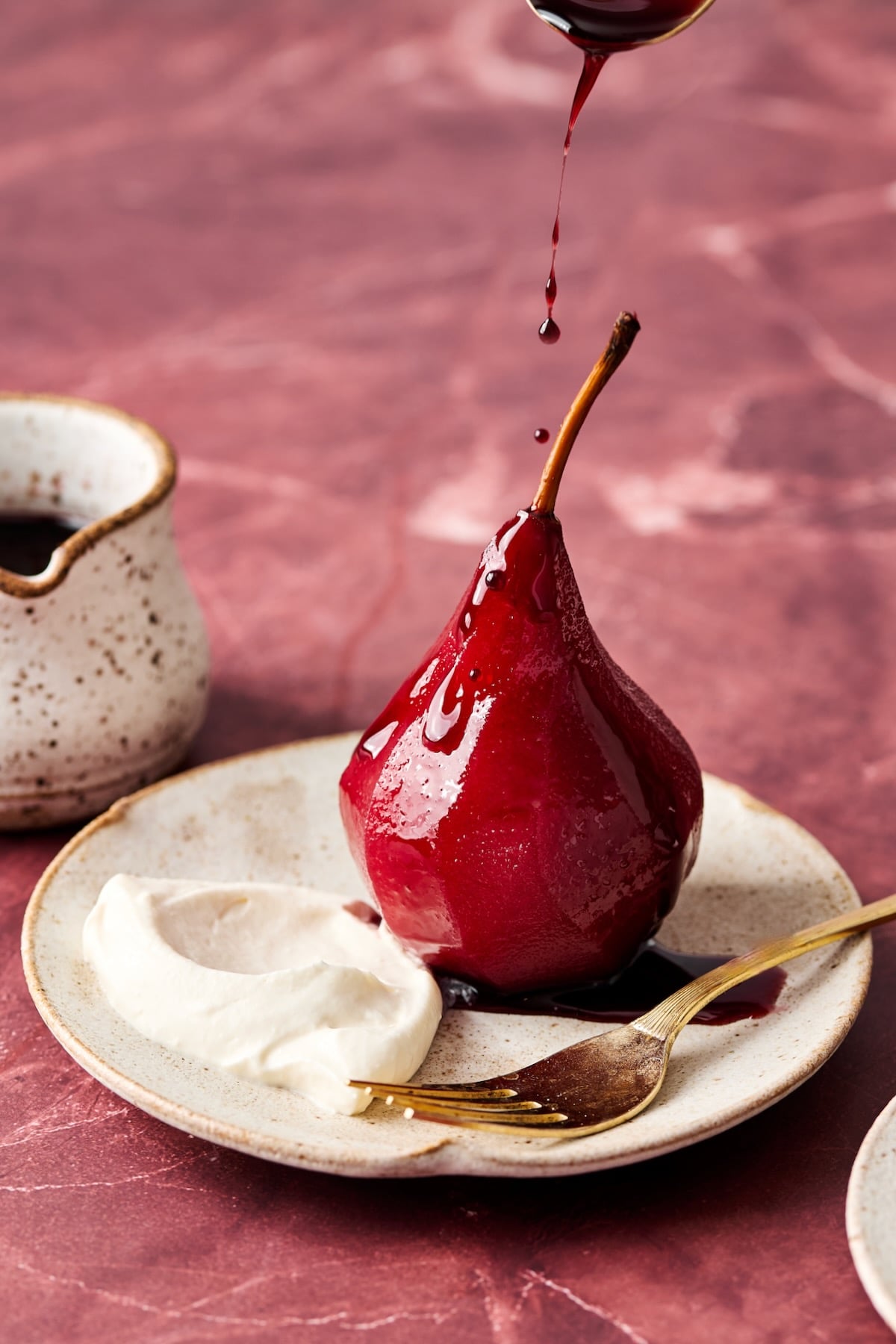 Red Wine Poached Pear with Mascarpone Whipped Cream on white plate with gold fork being drizzled with sauce.