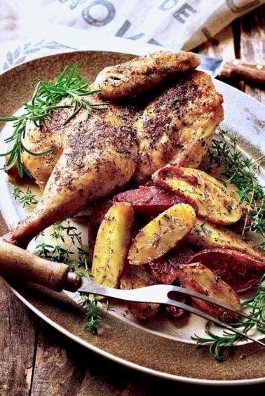 Roast Chicken Half with Fingerling Potatoes on gray-rimmed platter with. meat fork garnished with fresh rosemary and thyme.