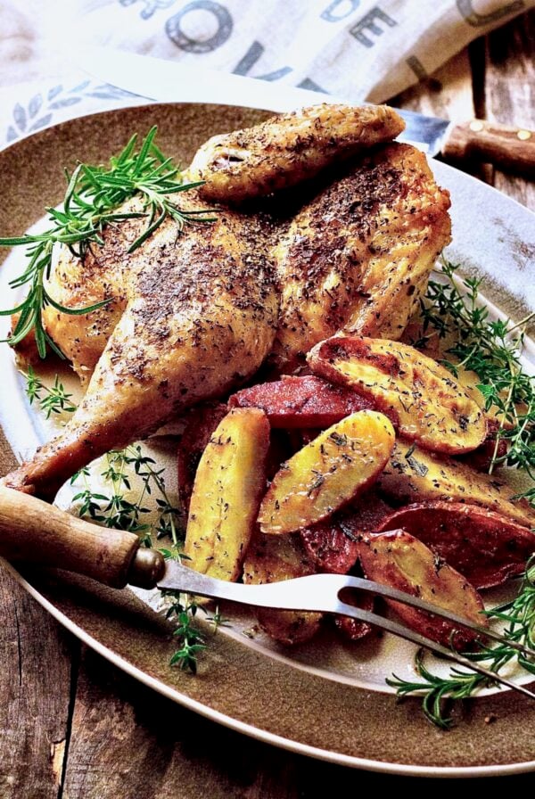 Roast Chicken Half with Fingerling Potatoes on gray-rimmed platter with. meat fork garnished with fresh rosemary and thyme.
