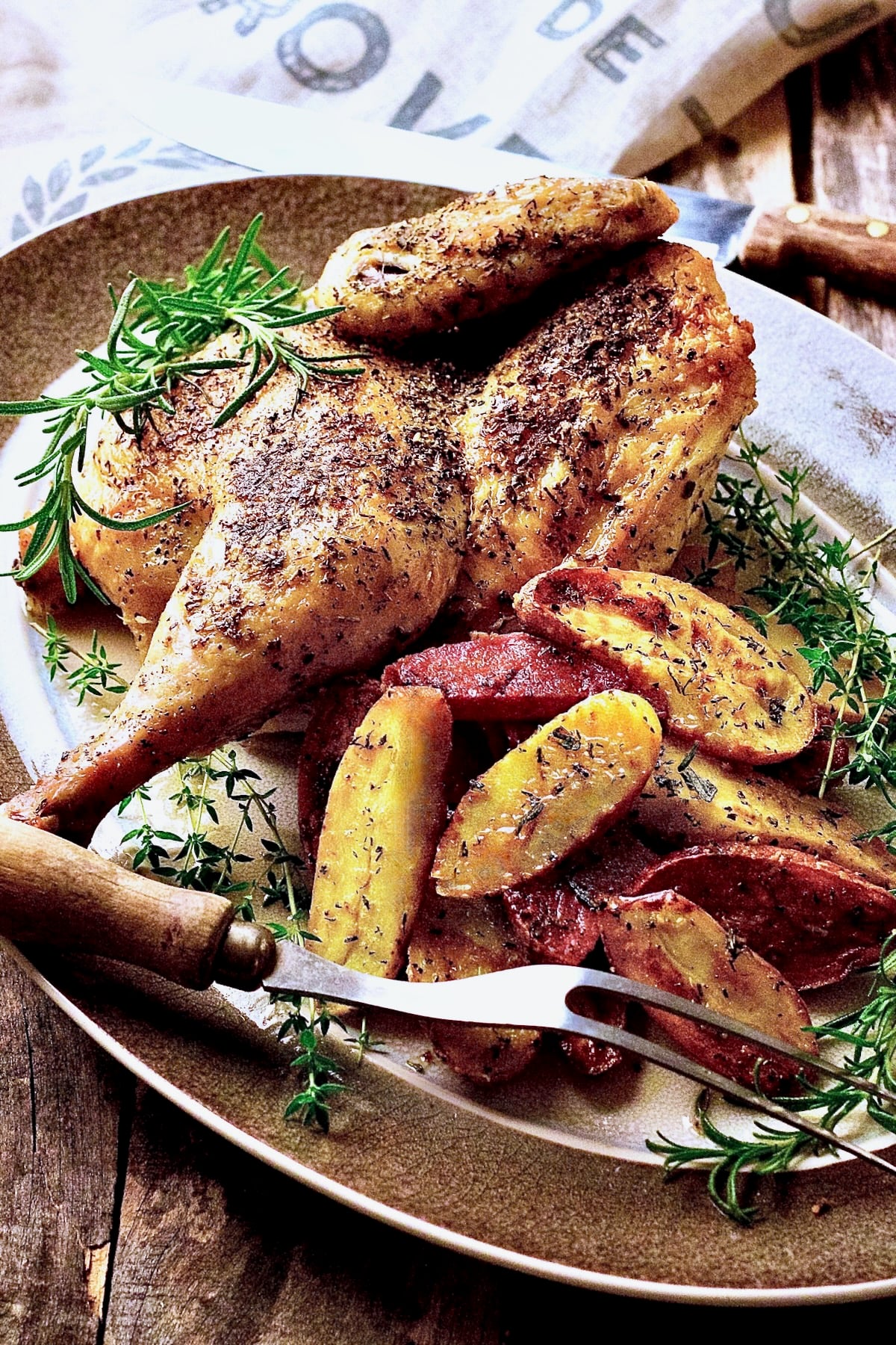 Roast Chicken Half with Fingerling Potatoes on gray-rimmed platter with. meat fork garnished with fresh rosemary and thyme.