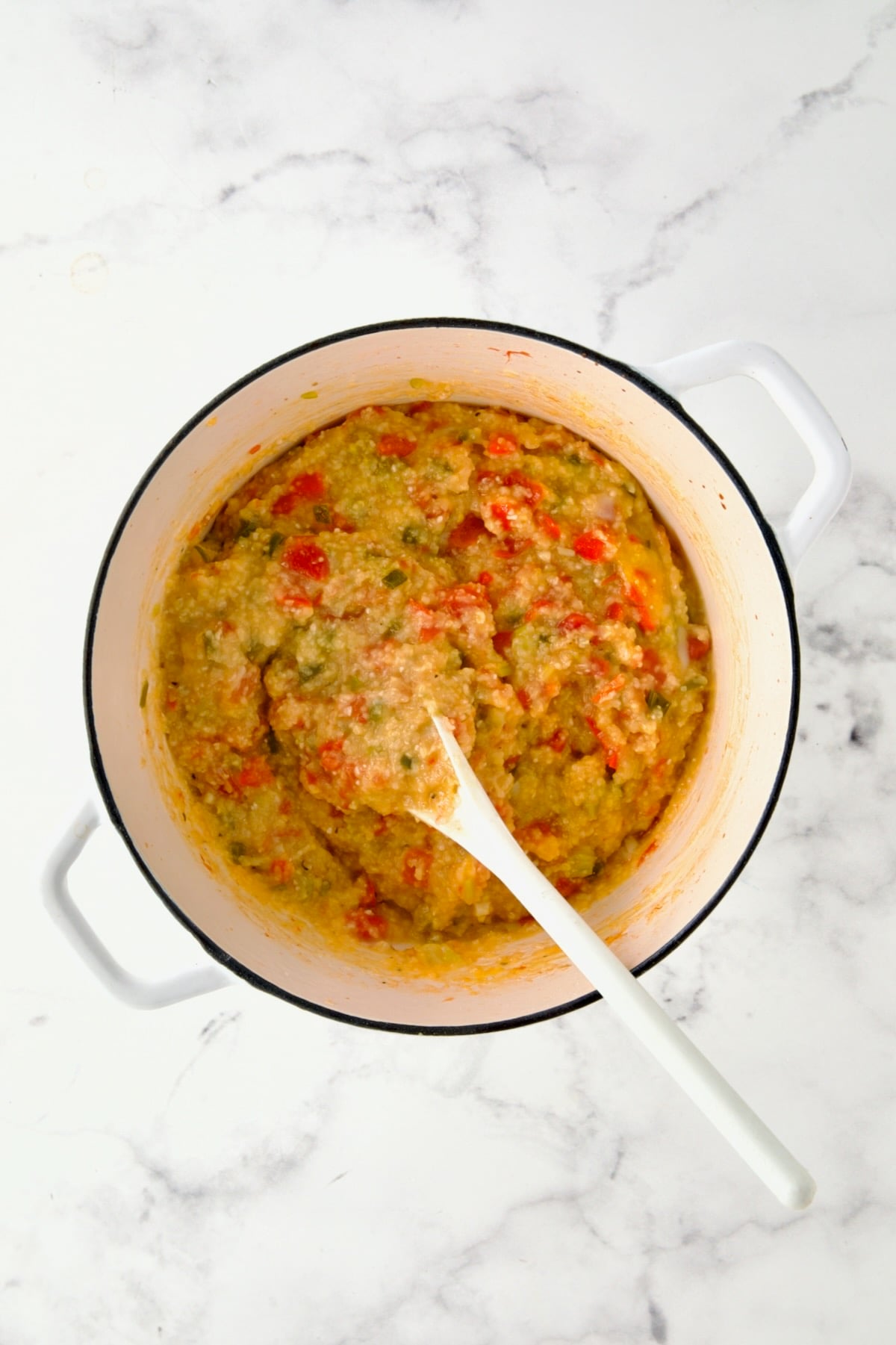 Egg being stirred into vegetable and grits combination with white mixing spoon in white Dutch oven.