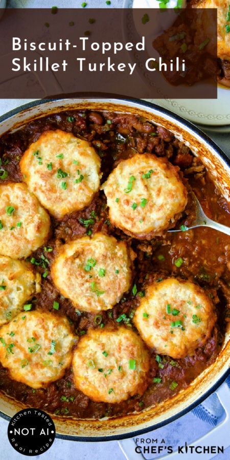 Pinterest graphic for Biscuit-Topped Turkey Skillet Chili showing it in a white enamel cast iron skillet with serving spoon.