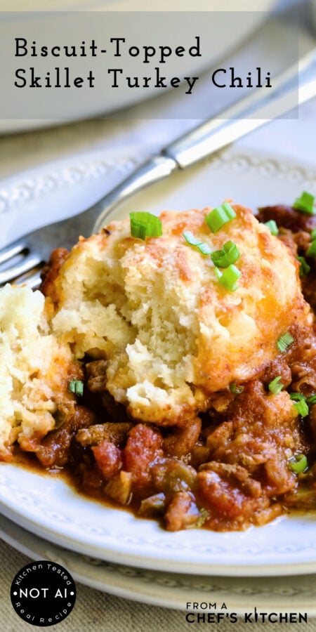 Pinterest graphic for Biscuit-Topped Turkey Skillet Chili showing it in a white enamel cast iron skillet with serving spoon.