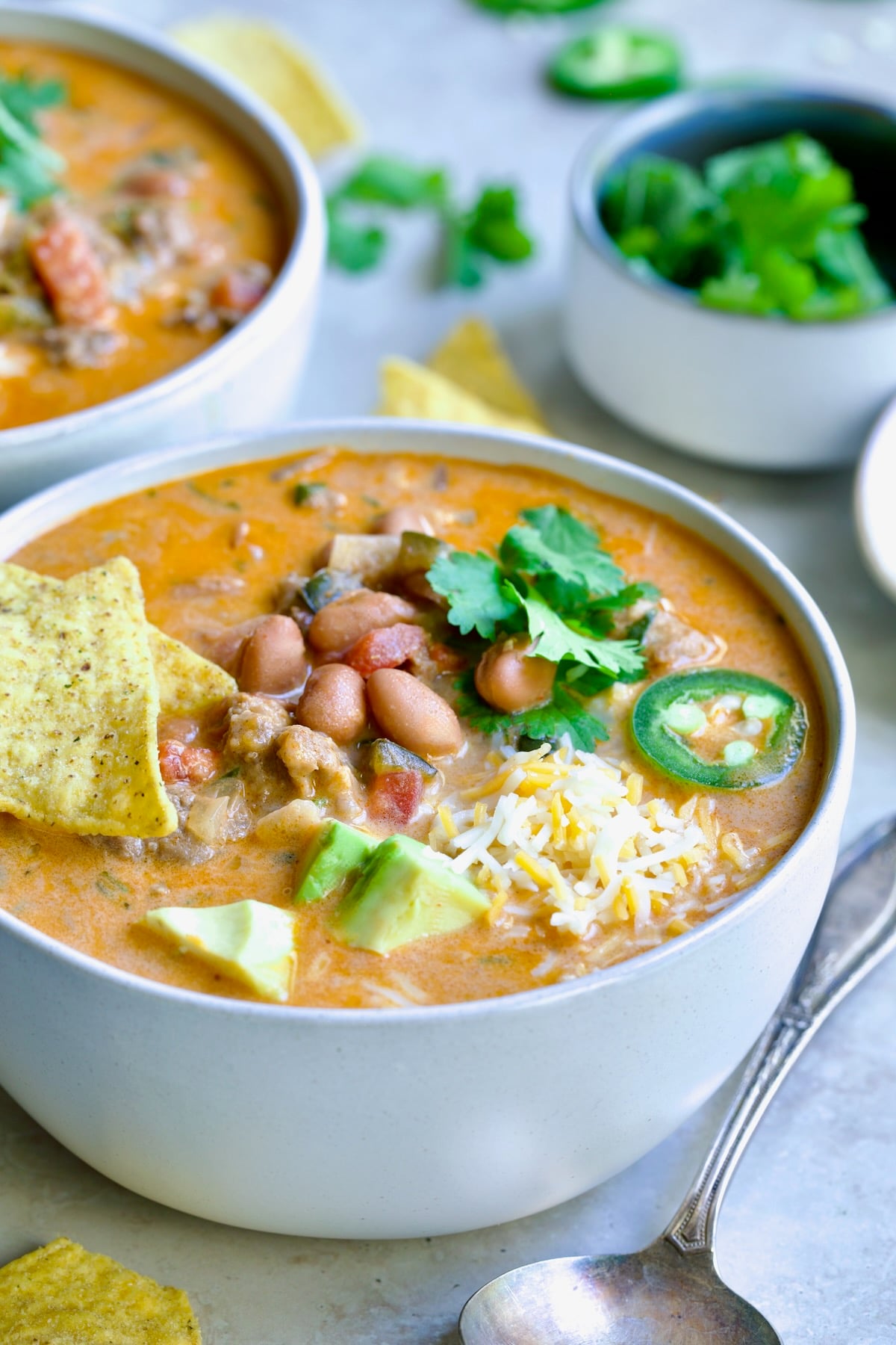One serving of Creamy Taco Soup in white bowl garnished with tortilla chips, avocado, cheese and jalapeno slices.