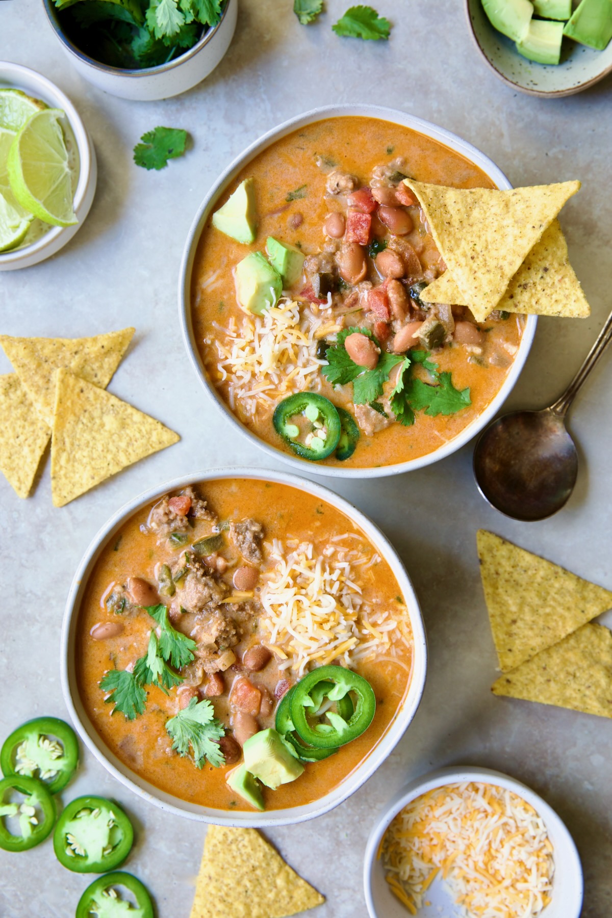 Two servings of Creamy Taco Soup in white bowls garnished with tortilla chips, avocado, cheese and jalapeno slices.