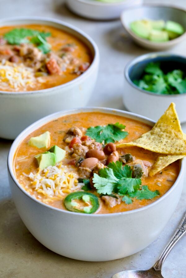 One serving of Creamy Taco Soup in white bowl garnished with tortilla chips, avocado, cheese and jalapeno slices.
