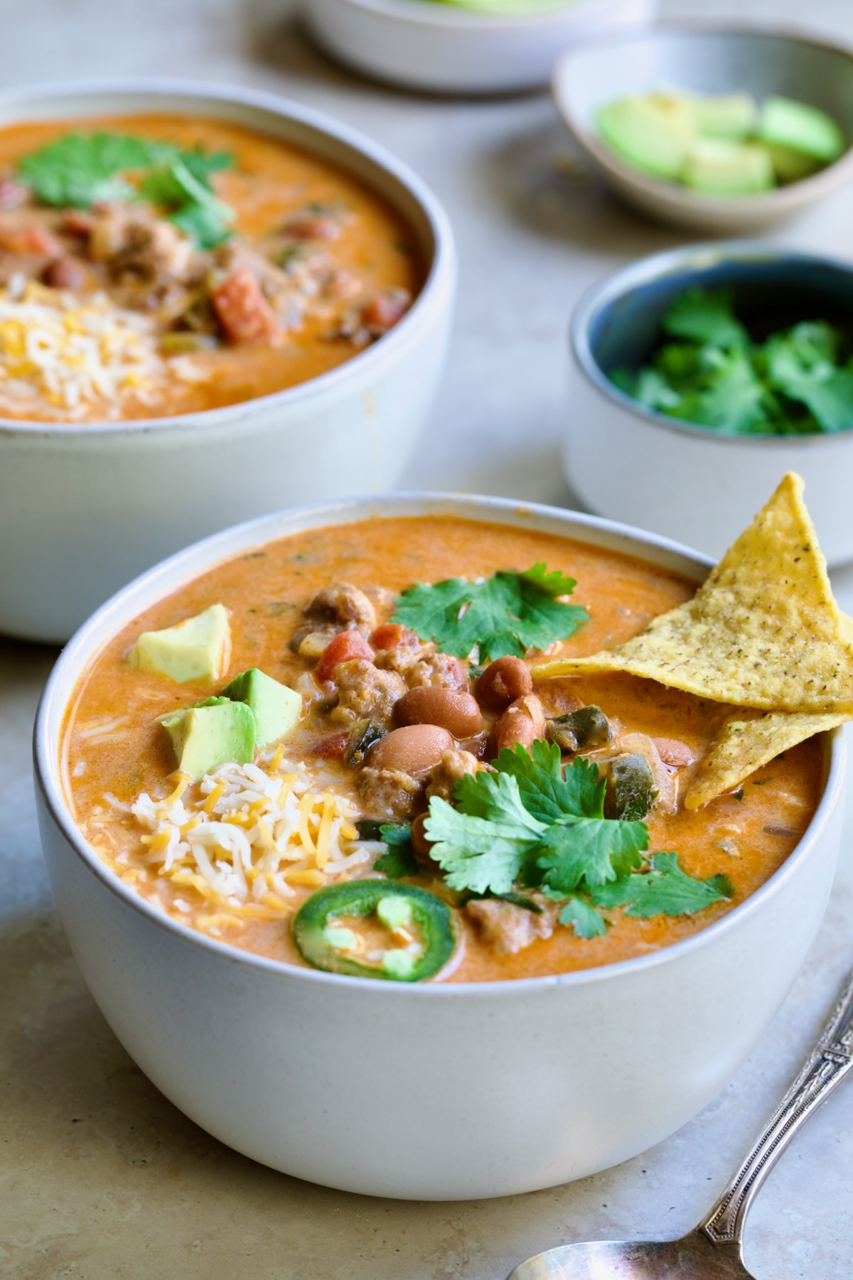 One serving of Creamy Taco Soup in white bowl garnished with tortilla chips, avocado, cheese and jalapeno slices.