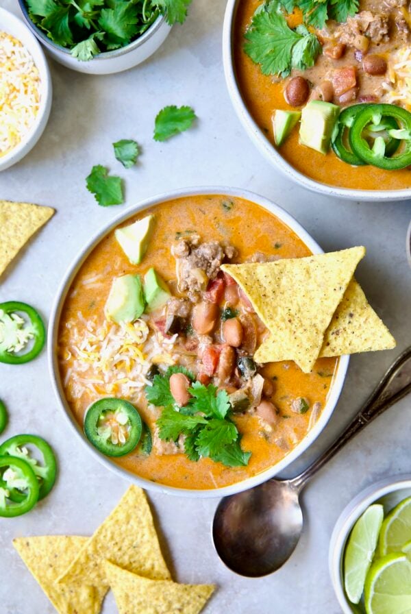 One serving of Creamy Taco Soup in white bowl garnished with tortilla chips, avocado, cheese and jalapeno slices.
