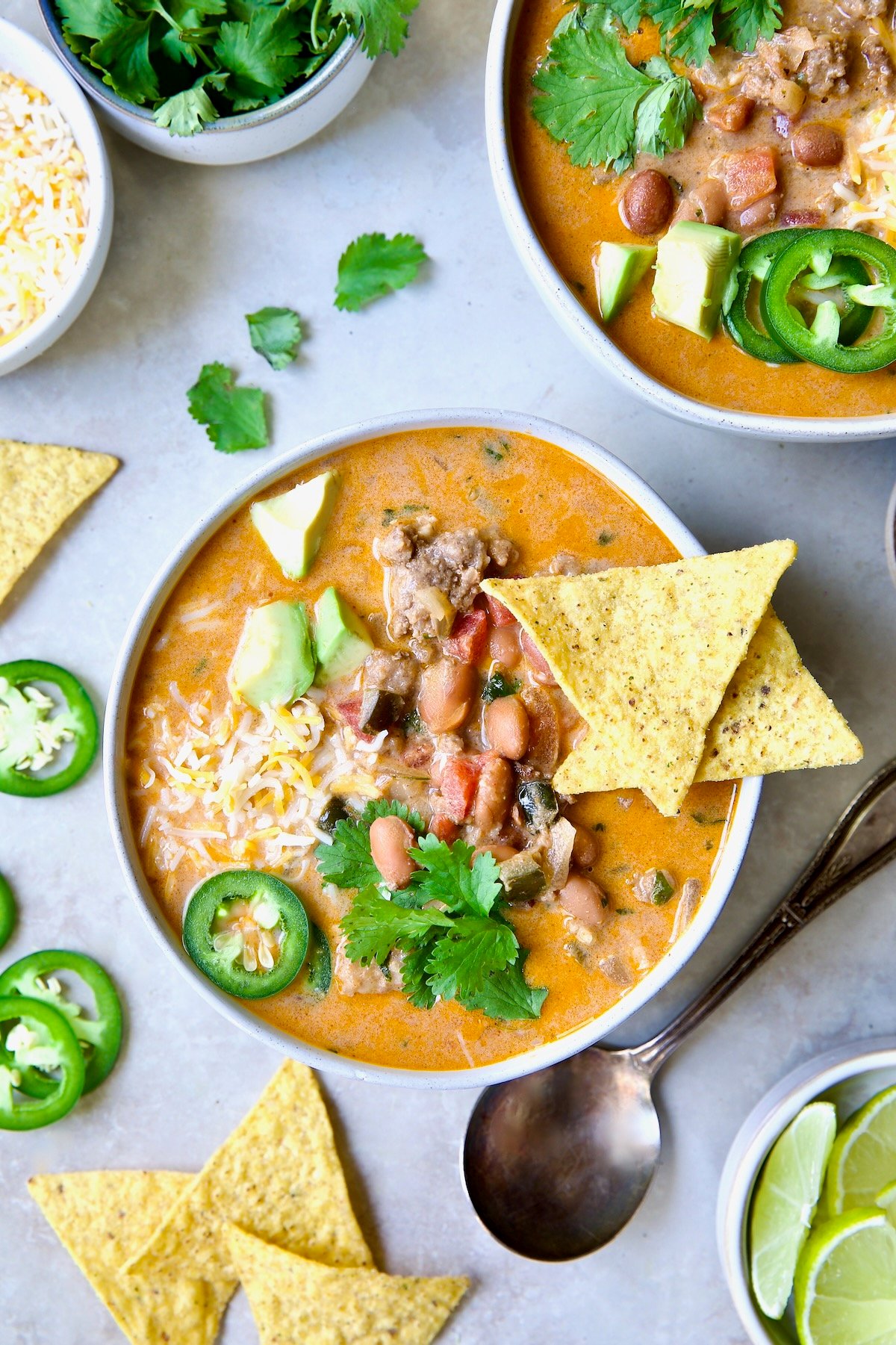 One serving of Creamy Taco Soup in white bowl garnished with tortilla chips, avocado, cheese and jalapeno slices.