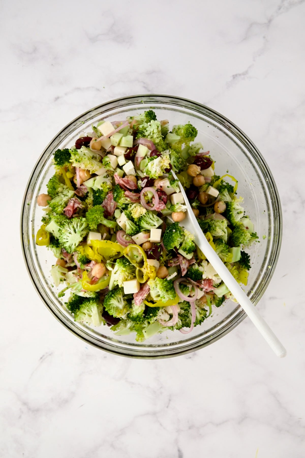Ingredients for Italian Broccoli Salad being mixed together in glass bowl with white mixing spoon.