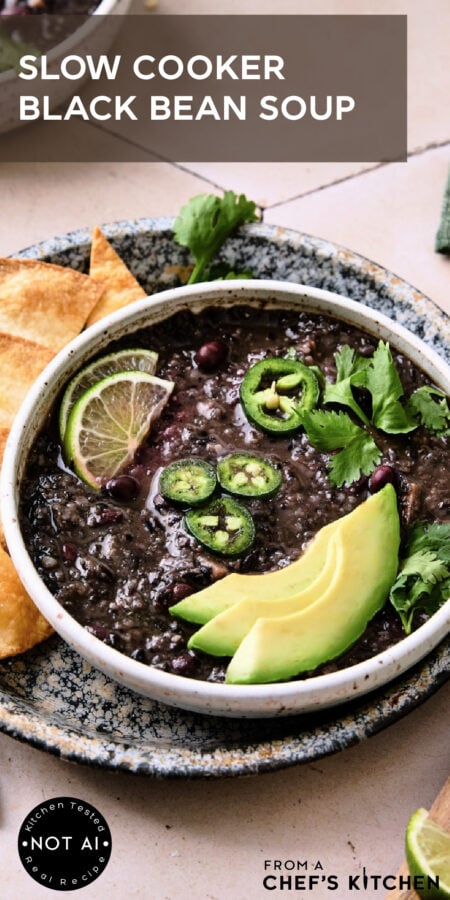 Pinterest graphic for Slow Cooker Black Bean Soup showing it in a white bowl garnished with lime wedges, jalapeno slices, cilantro and avocado.