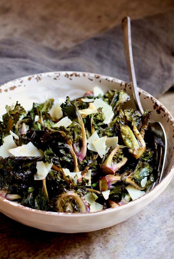 Roasted kale sprouts with shaved Parmesan cheese in white bowl with serving fork.