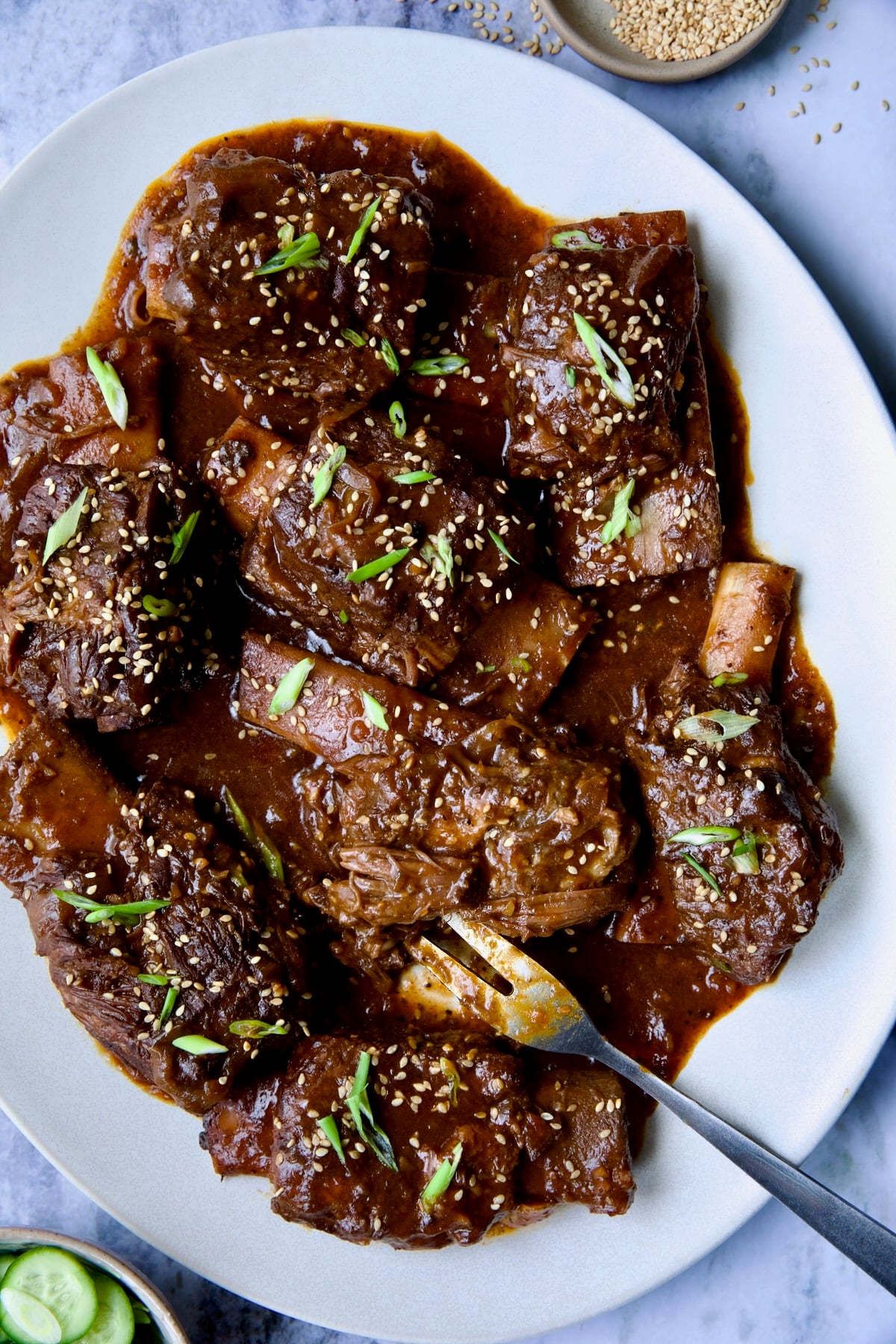 Korean Braised Beef Short Ribs on oval white platter with meat fork sprinkled with sesame seeds and thinly sliced scallions.